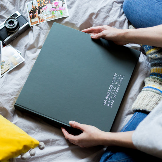 a black large wedding album is being held in a woman's hands whilst she is sitting on her bed. The album has been printed along the bottom in a simple, sharp contemporary font 