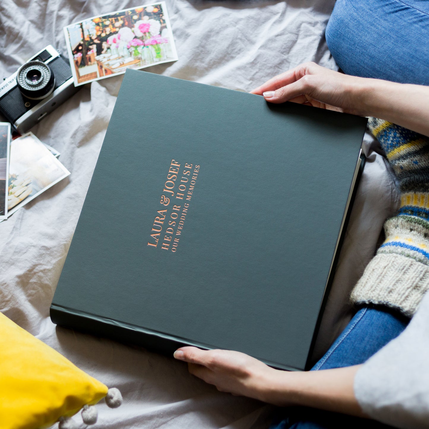 someone is sitting on a bed holding a large black wedding photo album . The wedding photo album has been printed on the front with a bride and groom's wedding details in a copper foil. There is also a camera and a set of photographs on the bed 