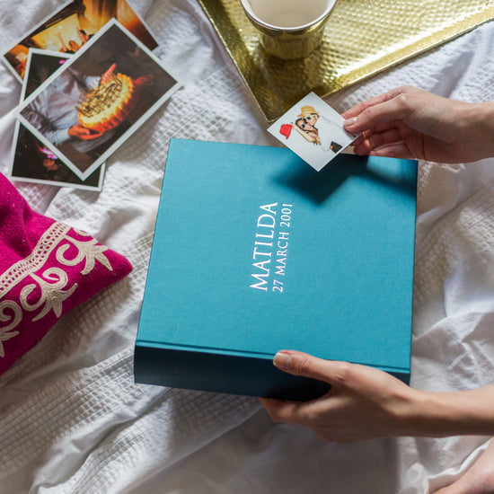 a woman is holding a blue photo album which has been personalised with a girl's name and birthday on the front. In the background are more photographs for the photo album