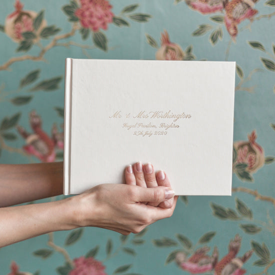 an ivory wedding guest book is being held up against a flowery wallpaper. The wedding guest book has been printed on the front with a calligraphy font in gold foil