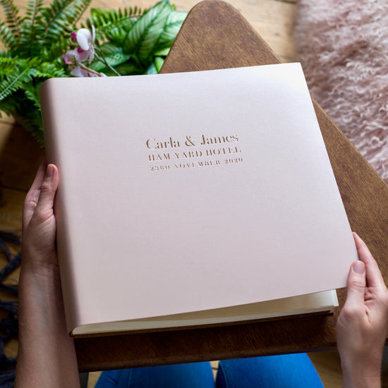on top of the wooden table is a big pale pink leather wedding album. It has been printed with wedding details on the front in gold foil