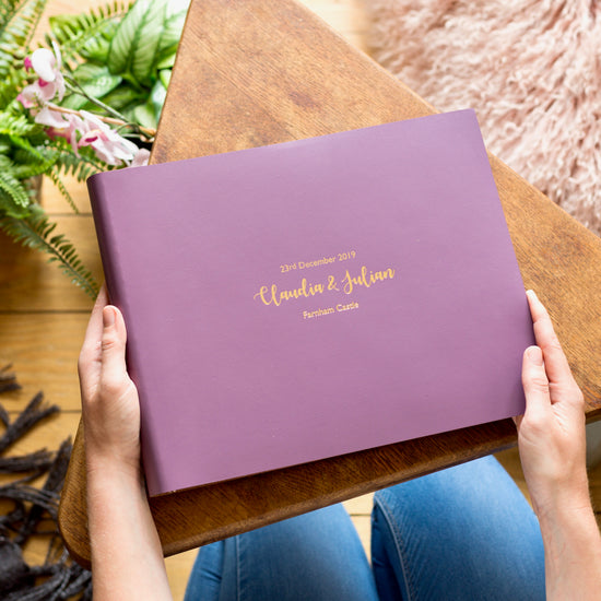 on a triangular wooden table is a purple wedding guest book that has been printed with gold wording. A woman is holding it in her hands