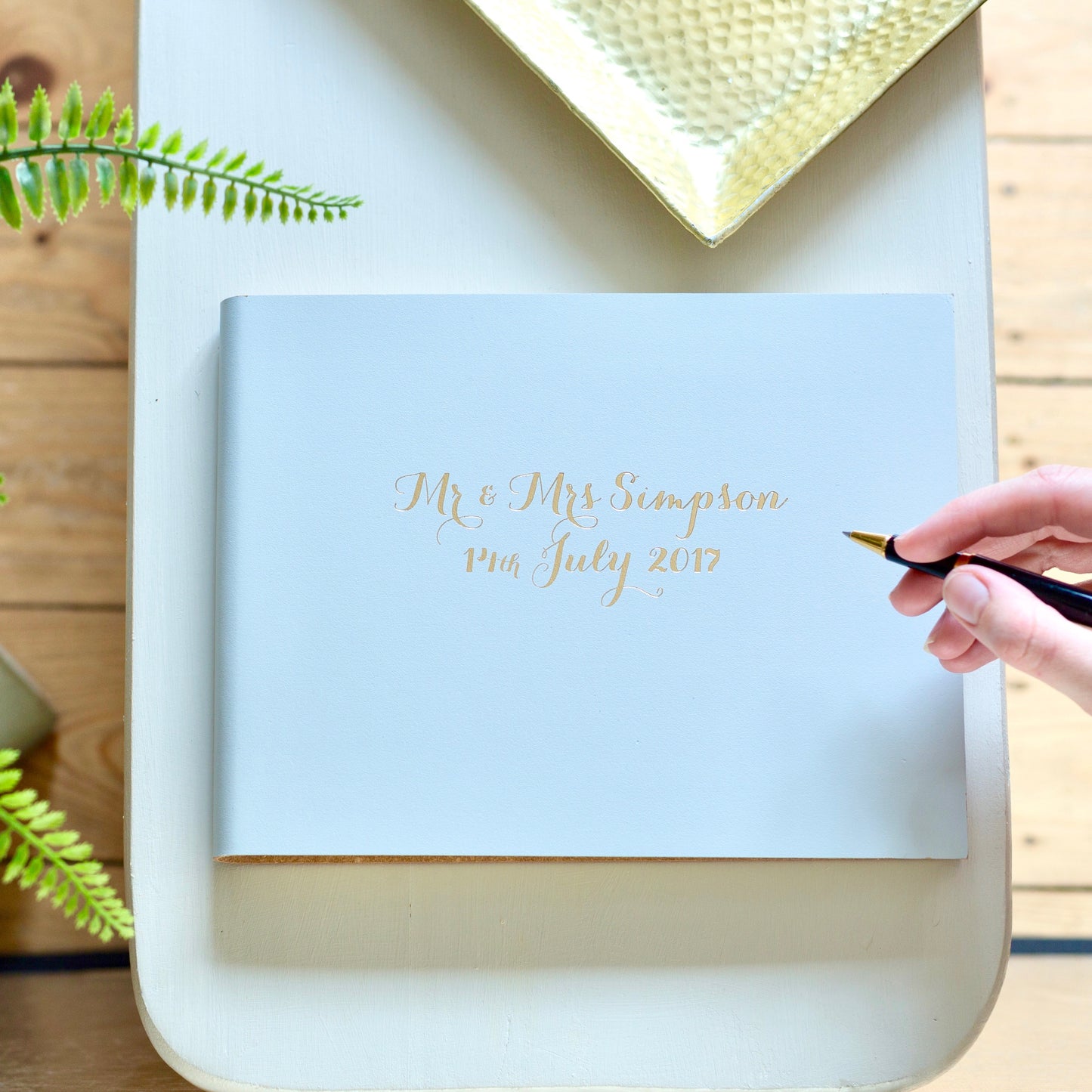 a wedding guest book is on a white table and someone has a pen in their hand poised to write a message in it. The book is grey and and has the names of thebride and groom on the front and the date of the wedding 
