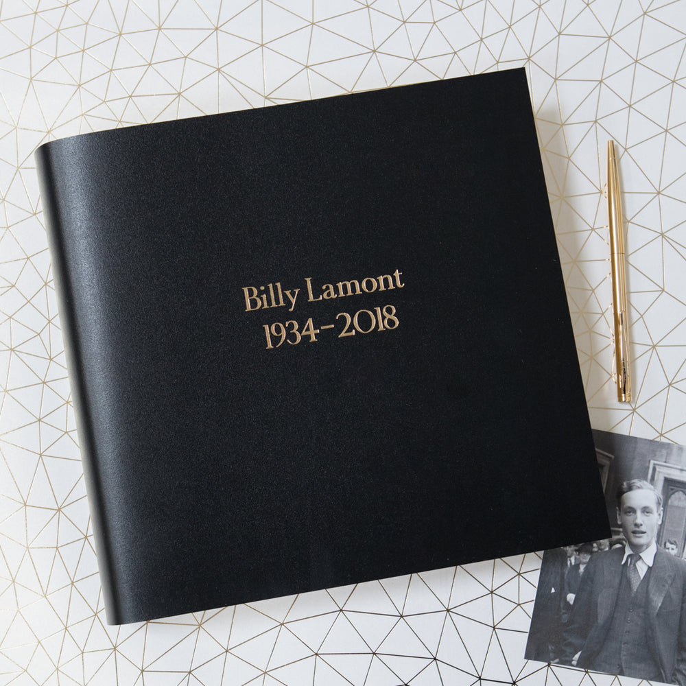 
                      
                        A black square condolence book has been printed with the name of a deceased man in gold lettering. There is also a gold pen by the side.
                      
                    