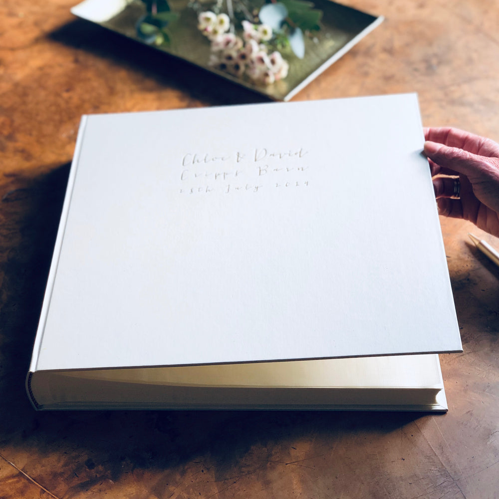 
                      
                        On a wooden table there is a large ivory wedding album that has been personalised with wedding details. 
                      
                    