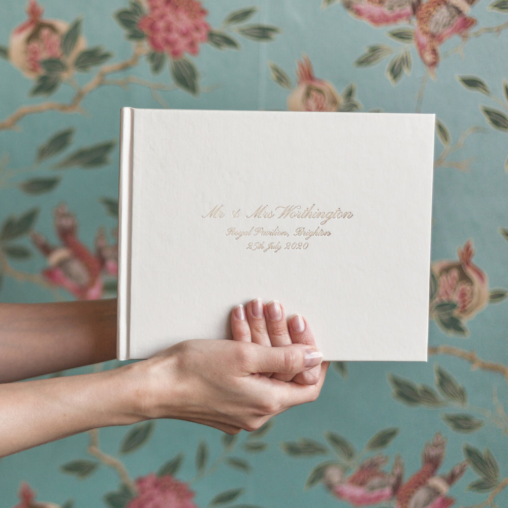An ivory wedding guest book is being held up against a flowery wallpaper. The wedding guest book has been printed on the front with a calligraphy font in gold foil.