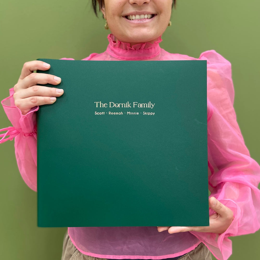 A woman in a pink top is holding a large green family album. Their family names have been included in the personalisation, which is with gold hot foil. 