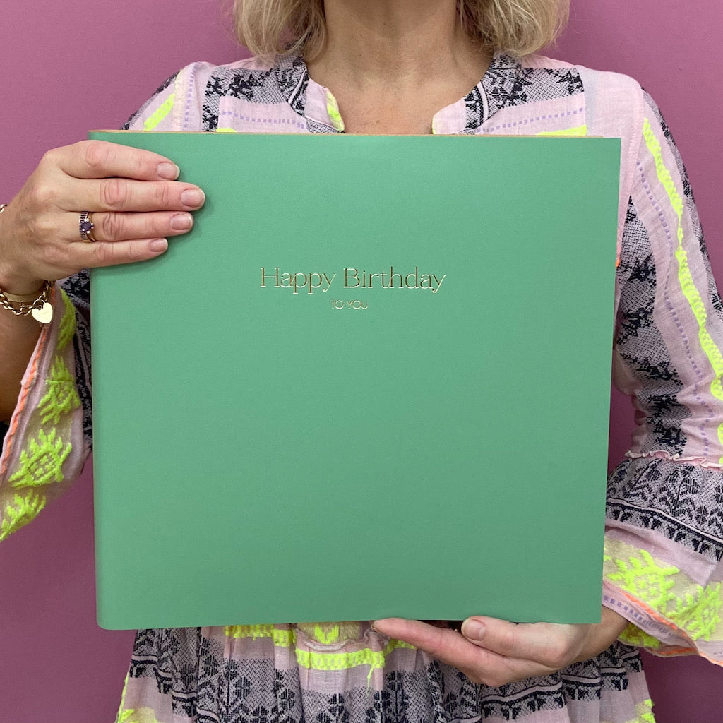 A woman in a pink dress is holding up a large green photo album. The photo album has been printed with the words Happy Birthday to you.