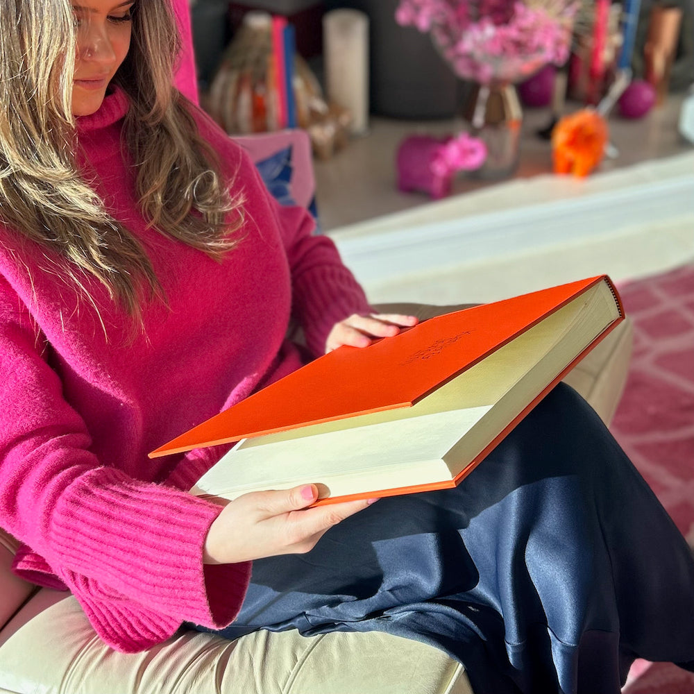 
                      
                        A young woman is sitting on a chair holding a large orange holiday photo album. She is looking at her holiday photos.
                      
                    