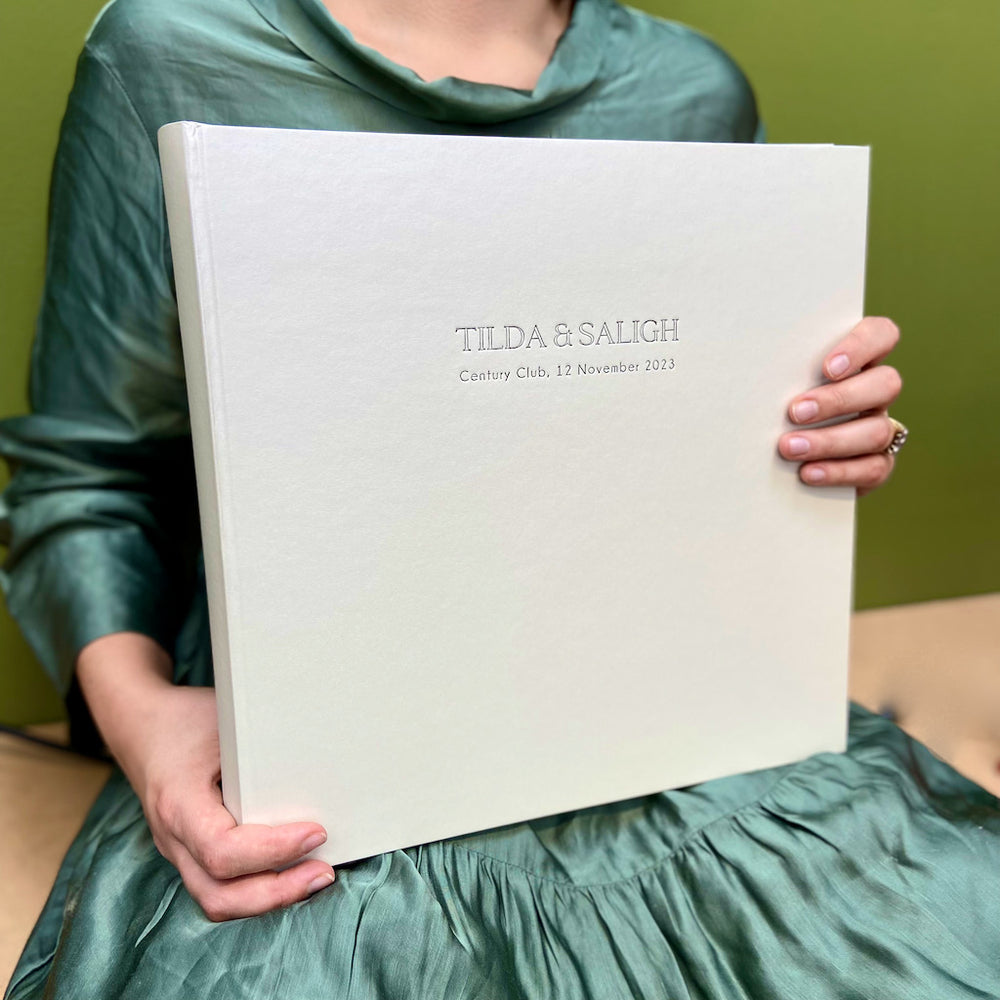 
                      
                        There is a woman in a green dress holding up a large white jewel album. It has been personalised with someone's first names, a location and has also been dated. 
                      
                    