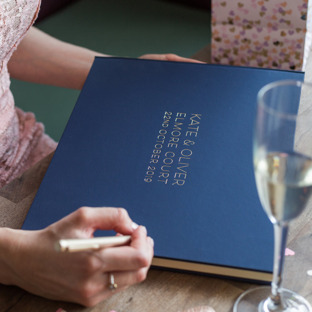 
                      
                        There is a large landscape jewel album which has been personalised with gold hot foil. A woman has a pen in her hand  ready to write a message. 
                      
                    