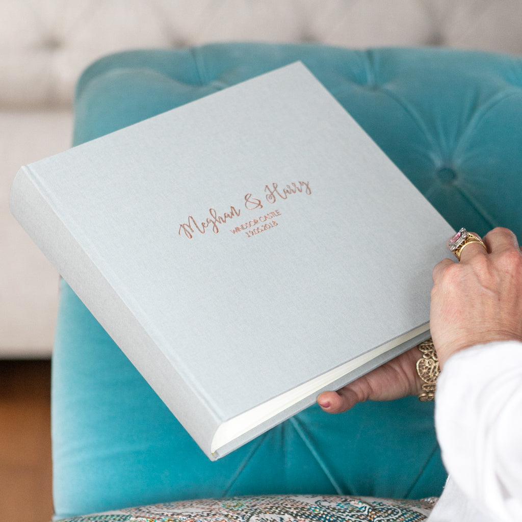 There is a woman holding a large linen wedding guest book. She is about to open the book whilst sitting on a blue sofa. 