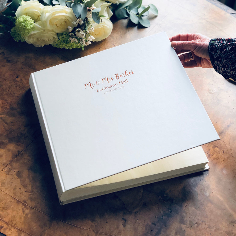 
                      
                        On a table there is a big bunch of flowers and a large square wedding album. The wedding album has been personalised with wedding details on the front.
                      
                    
