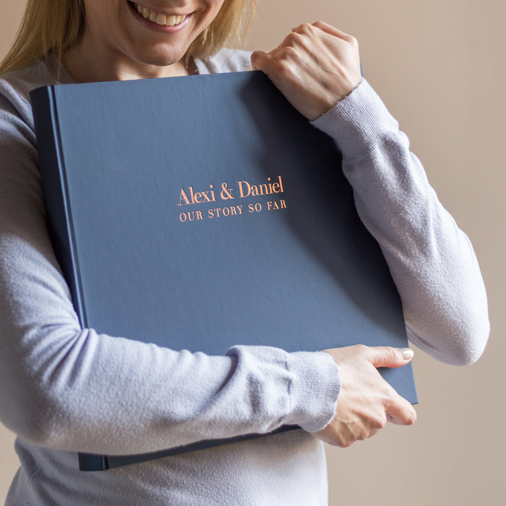 
                      
                        A woman is smiling whilst holding her wedding album to show her friends. Her and her husbands names are printed on the front in copper hot foil.
                      
                    