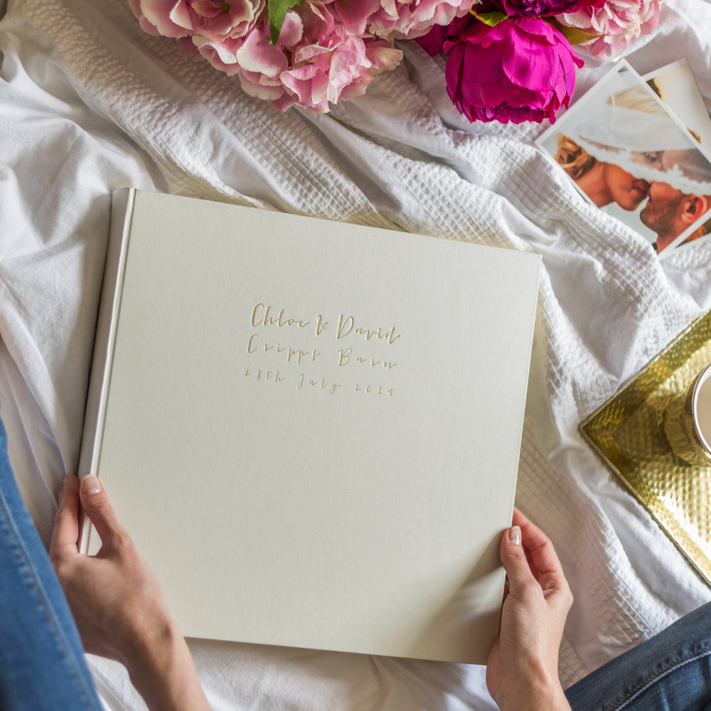 A large wedding photo album is on a white bed sheet and someone is holding it in their hands. There are also some wedding photos in the frame.