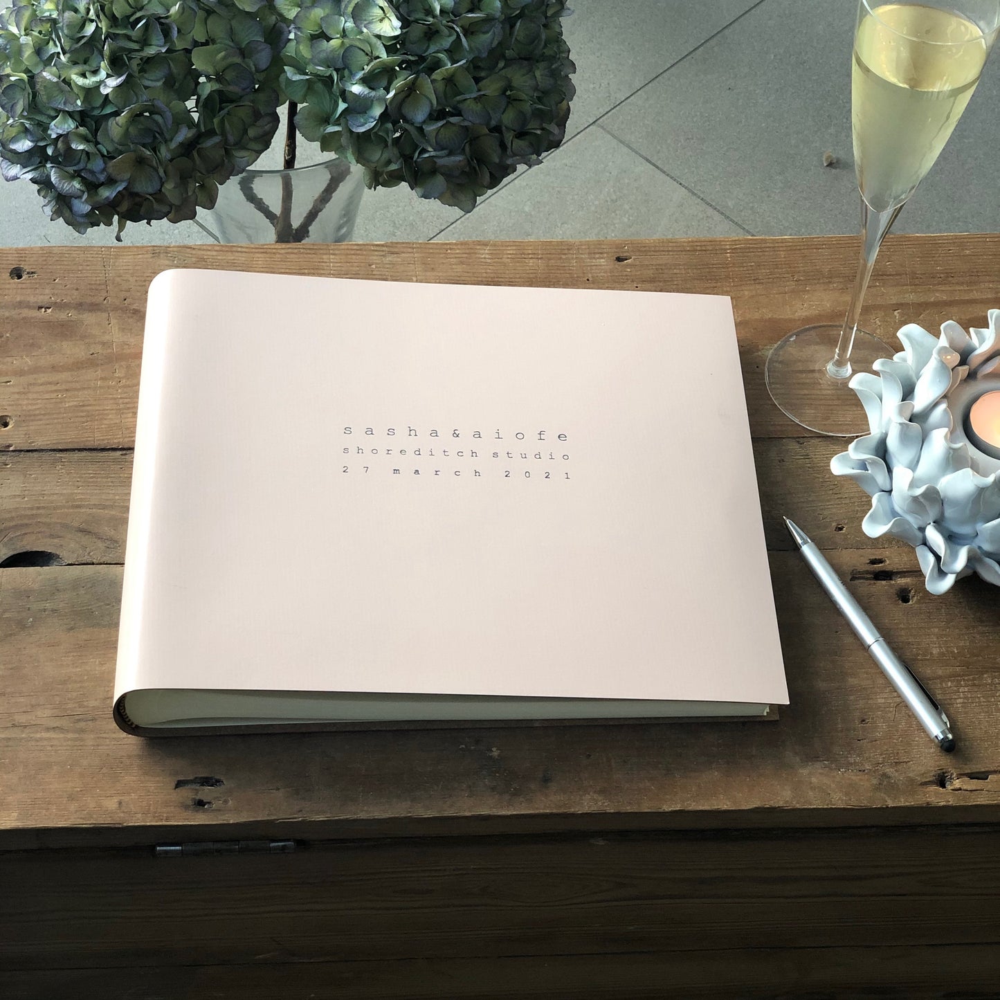 There is a large leather photo album placed on a wooden table. A glass of champagne and a lit candle is also in the photograph. This beautiful album has been personalised with details on the front. 