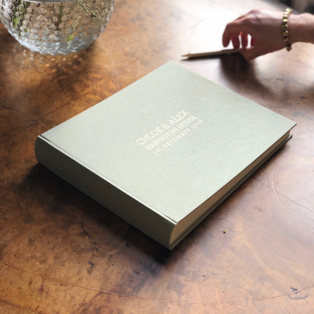 
                      
                        On a walnut table lies a wedding guest book in the shade Eau de Nil. A woman has a pen in her hand and she is about to write in the book. The guest book has a personalisation on the front.
                      
                    