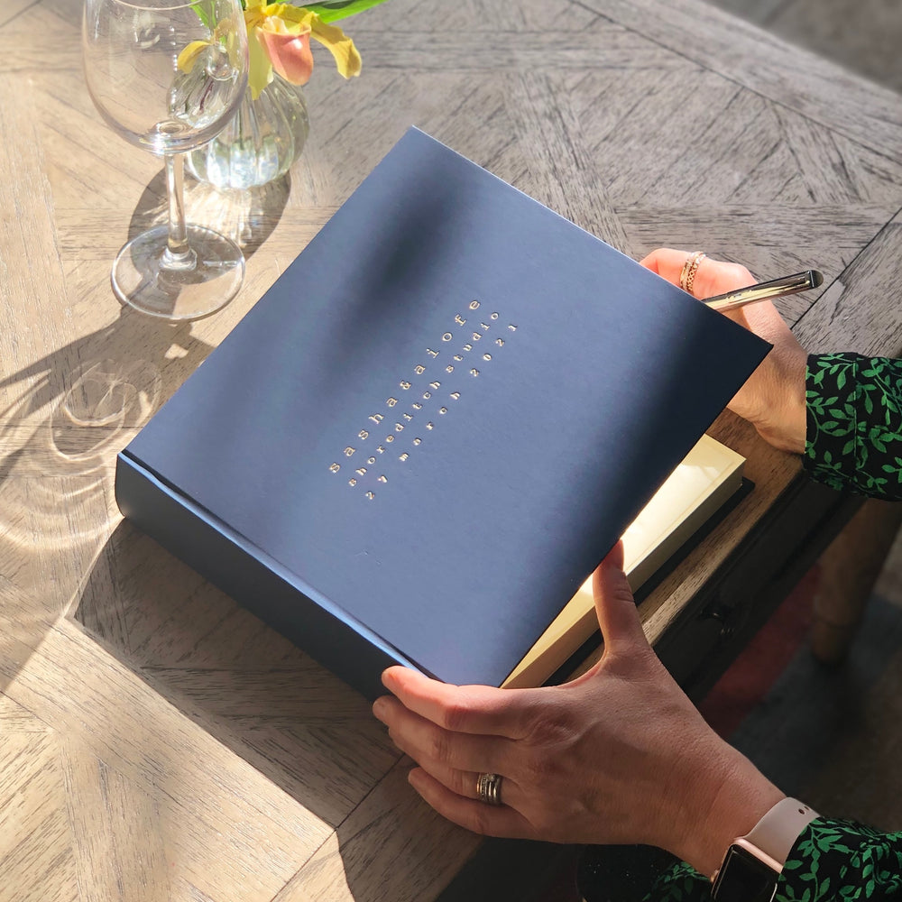 There is a woman poised with a pen in her hand. She is about to write a message in a wedding album which has personalised details on the front. There is also a posy of flowers and a wine glass on the wooden table.