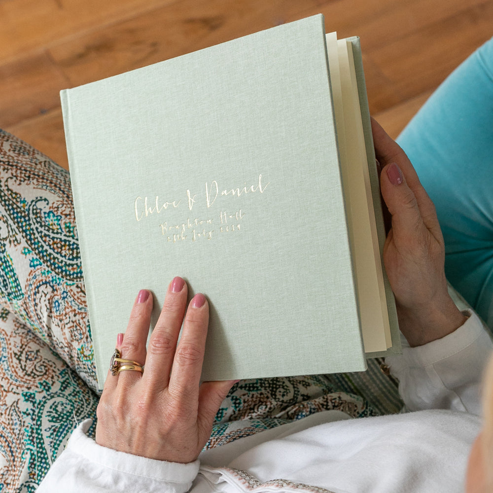
                      
                        A woman is sat on a blue sofa opening up her album. The album has been personalised on the front with gold hot foil.
                      
                    