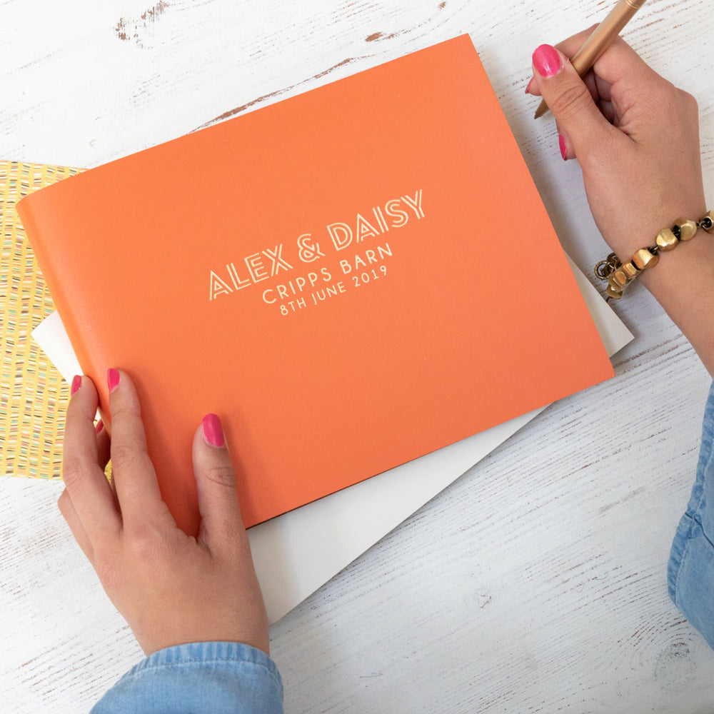 
                      
                        There is an orange landscape guest book with wedding details printed on the front in hot foil. She is holding a pencil ready to write a message inside. 
                      
                    